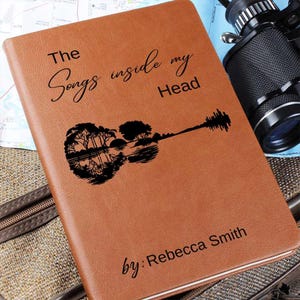 May include: A brown leather journal with the text "The Songs inside my Head" and a silhouette of a guitar with trees reflected in the body. The text "by: Rebecca Smith" is at the bottom of the journal.