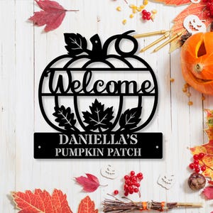 May include: Black metal pumpkin-shaped sign with the word "Welcome" cut out. Below, a rectangular plaque reads "DANIELA'S PUMPKIN PATCH". The sign is decorated with leaf cutouts, perfect for fall decor.