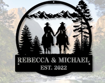 May include: A circular black metal wall sign featuring a silhouette of two people riding horses through a forest with mountains in the background. The sign includes the text REBECCA & MICHAEL and EST. 2022 cut out in the lower rectangular section.
