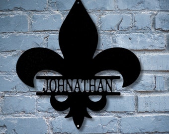 May include: Black metal fleur-de-lis wall art with the name "JOHNATHAN" cut out in the center. The decorative piece is mounted on a blue brick wall. The fleur-de-lis has a classic three-petal design.