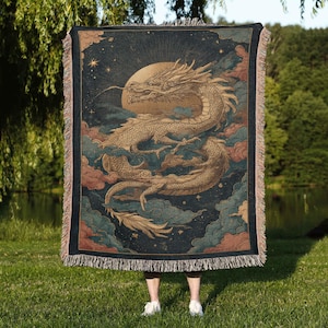 A person is standing in a grassy area, holding a large tapestry or blanket featuring a dragon and a starry night sky.