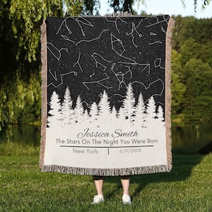 May include: A black and white woven blanket featuring a starry night sky design with constellations. The bottom of the blanket has a white forest silhouette and the text "Jessica Smith, The Stars On The Night You Were Born, New York, 6/9/2005."