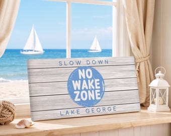 May include: A rectangular wooden sign with the text "SLOW DOWN" above a blue circle that reads "NO WAKE ZONE LAKE GEORGE". The sign is placed on a windowsill with a view of the ocean and sailboats.