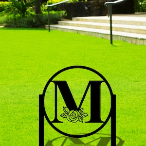 May include: Black metal garden stake with a circular design featuring the letter "M" and a stylized rose. The stake is placed in a green grassy lawn.