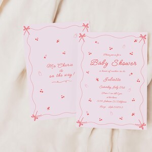 May include: Pink baby shower invitation with a red border and red illustrations of cherries and bows. The text reads "Please join us for a Baby Shower in honor of mother-to-be Juliette. Saturday, July 21st. 2-5 pm at her home. 1234 Johnson St. California. RSVP 555-123-4567."