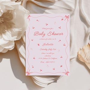 May include: Pink baby shower invitation with a red string border and red illustrations of cherries, pacifiers, and a rattle. The text reads "Please join us for a Baby Shower in honor of mother-to-be Juliette, Saturday July 21st, from 11 am to 4 pm, at her home, 1234 Johnson St. California, RSVP 555-098793."