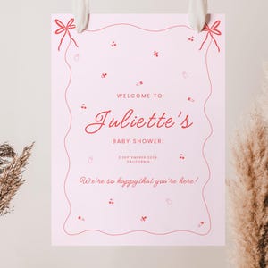 May include: A pink welcome sign with a wavy border and red text that reads "Welcome to Juliette's Baby Shower! September 2024, California. We're so happy that you're here!"