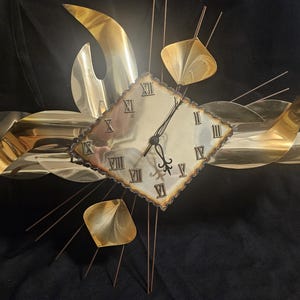 May include: A gold-toned metal wall clock with a diamond-shaped face and Roman numerals. The clock is surrounded by abstract, curved metal shapes and thin rods, creating a mid-century modern design. The clock hands are black.