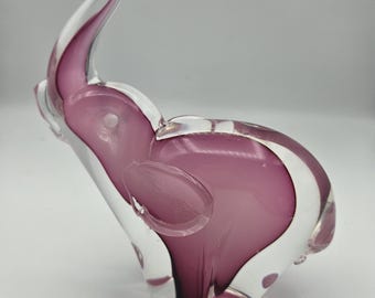 May include: A decorative glass elephant figurine. The elephant is a translucent clear glass with a vibrant purple interior. The trunk is curved upwards, and the ears are flared. The figurine is a unique home decor item.