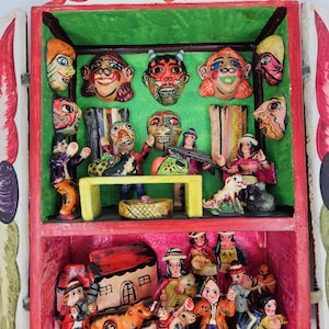 Puede incluir: Una colorida caja de sombras de arte popular hecha a mano con múltiples niveles. El nivel superior presenta caras de máscaras pintadas. El nivel inferior representa figuras en miniatura, animales y una casa. La caja está pintada de rojo, verde y rosa.
