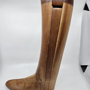 May include: A vintage wooden boot stretcher, with a foot-shaped base and an adjustable shaft. The wood has a warm, natural tone with darker accents. The top section is marked with the letter "L".