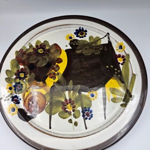 Pode incluir: Um prato de cerâmica branco com uma borda marrom. O prato apresenta um desenho pintado à mão de uma figura marrom estilizada com acentos florais amarelos, verdes, azuis e vermelhos.