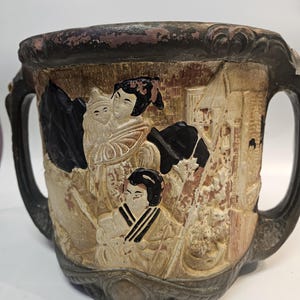 May include: A detailed, antique ceramic cup with two handles. The cup features a relief design depicting figures in traditional clothing, with black, beige, and brown tones. The rim and handles are dark brown, and the base has a decorative pattern.