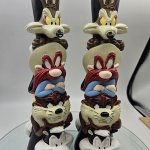 May include: A pair of ceramic figurines featuring characters from the Looney Tunes cartoon. The figurines are stacked on top of each other, with the top character being Wile E. Coyote, followed by Yosemite Sam, Taz, and finally, Pepe Le Pew. The figurines are painted in bright colors and have a cartoonish style.