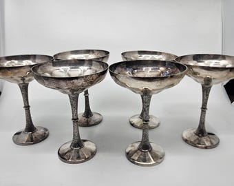 May include: Six vintage silver champagne coupe glasses. Each glass has a wide, shallow bowl and a long, ornate stem. The glasses are arranged on a white surface, showcasing their reflective surfaces and classic design.