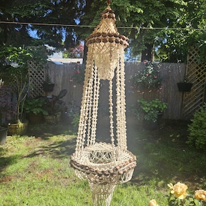May include: A hanging macrame plant holder with a tiered design. The holder is made of off-white macrame and adorned with brown shells. It is suspended from a hook and hangs in a garden setting with green grass and foliage.