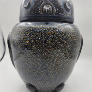 Könnte beinhalten: Eine schwarze Keramik-Dose in Form einer Eule mit blauen und goldenen Maldetails. Die Dose hat einen runden Körper mit einer strukturierten Oberfläche und einen Deckel mit einem erhabenen Eulendesign.