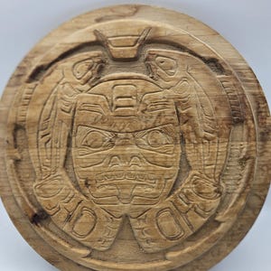 May include: A wooden carving of a face with intricate details. The carving is circular and has a raised border. The face is surrounded by two figures, possibly birds, with their wings outstretched.