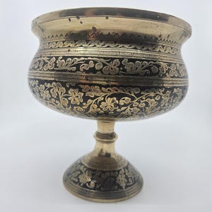 May include: A gold-coloured metal bowl with a pedestal base. The bowl features intricate floral patterns in black against a gold background. The rim and base also have decorative detailing. The bowl is likely used for display or serving.