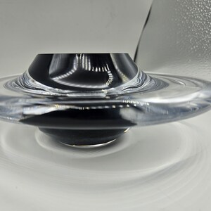 May include: A clear glass bowl with a black centre, resembling a flying saucer. The bowl has a smooth, rounded edge and a reflective surface.
