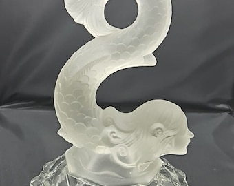 May include: A clear glass candlestick with a mermaid design. The mermaid is curled into an 'S' shape with her tail forming the candle holder. The base is a circular, scalloped design.