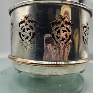 Puede incluir: Calienta-té de plata con una banda de cobre y diseños intrincados recortados. El calienta-té tiene tres patas y está sobre una superficie de vidrio.