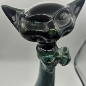 Puede incluir: Una figurita de cerámica de gato negro y verde con una expresión de guiño. El gato tiene un cuello largo y delgado y una cola que se enrosca alrededor de su cuerpo. La figurita está esmaltada en un color verde oscuro y negro con un toque de azul.
