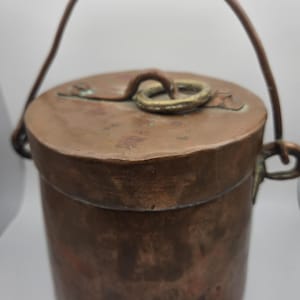 Puede incluir: Un pequeño recipiente redondo de cobre con tapa y asa. El recipiente tiene un aspecto desgastado y antiguo, probablemente sea antiguo.