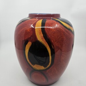 Könnte beinhalten: Eine dekorative Keramikvase mit glänzendem Finish. Die Vase ist hauptsächlich tiefrot, mit schwarzen, gelben und orangefarbenen abstrakten Mustern. Der Rand ist dunkelblau.