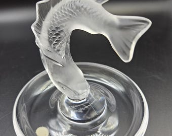 May include: A clear glass ring holder shaped like a fish. The fish is in a jumping pose with its tail raised. The ring holder is on a clear glass base.