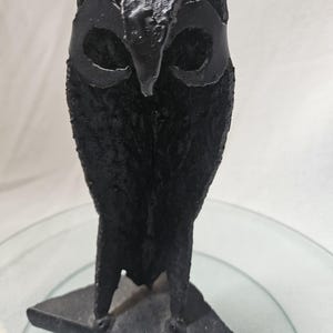 Puede incluir: Una escultura de búho de metal negro con ojos grandes. El búho está posado sobre una base triangular.