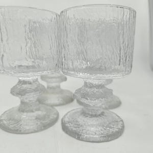 Pode incluir: Quatro taças de vidro transparente texturizado com haste curta e taça cilíndrica larga. O vidro tem uma textura áspera, semelhante à casca. As taças estão dispostas sobre uma superfície branca.