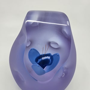 Puede incluir: Una escultura de vidrio artístico de color lavanda con un diseño de flor azul. La flor tiene múltiples capas de pétalos azules y estambres marrones. La escultura tiene una forma suave y redondeada con un acabado esmerilado.