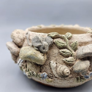 Può includere: Un piccolo vaso rotondo in terracotta con una superficie testurizzata. Il vaso è decorato con disegni in rilievo di funghi, foglie e lumache. Il vaso è di colore marrone chiaro con accenti verdi e bianchi.