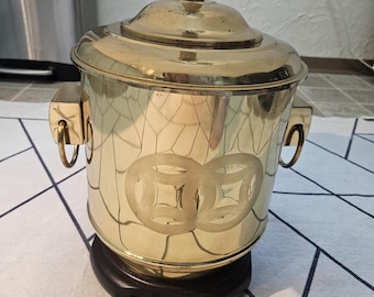 May include: A vintage brass lamp with a cylindrical body and a lid. The lamp features two decorative handles and a black wooden base. The body of the lamp has an etched design of two overlapping coins. The lamp is topped with a white lampshade.