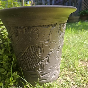 May include: A dark gray ceramic planter with intricate embossed floral and flame-like patterns. The wide-mouthed pot sits on a bed of green grass, showcasing its detailed design and textured surface. A decorative garden accent.