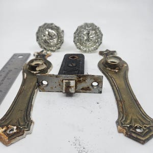Peut inclure: Deux poignées de porte en verre transparent, deux plaques de porte allongées en laiton à la finition usée et un mécanisme de serrure à mortaise en métal noir avec le mot Corbin visible. Une règle en acier inoxydable est sur la gauche.