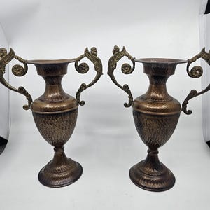 Könnte beinhalten: Ein Paar passende bronzefarbene Metallurnen mit einer texturierten, gehämmerten Oberfläche an Körper und Fuß. Jede Urne hat zwei verzierte, geschwungene Griffe, die mit kleinen skulptierten Figuren und Blumenmotiven verziert sind.