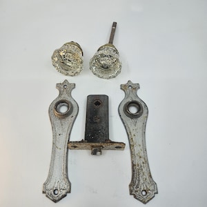 Peut inclure: Deux poignées de porte en verre transparent à facettes avec des bases dorées, deux plaques de propreté allongées en métal argenté patiné avec des détails décoratifs, et un mécanisme de verrouillage central en fer avec un trou carré.