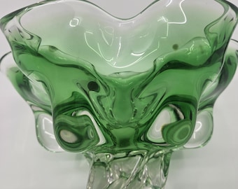 May include: A green glass bowl with a unique, wavy design. The bowl has a clear, twisted base and a flared, petal-like rim. The glass has a slight green tint, creating a vintage aesthetic. The bowl is likely used for decorative purposes.