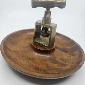 Puede incluir: Un cascanueces vintage con mecanismo y mango de latón, montado sobre una fuente ovalada de madera. El cascanueces presenta un mecanismo de tornillo para romper nueces. Se aprecia la veta de la madera del plato.