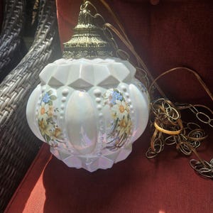 May include: A hanging pendant lamp with a spherical, iridescent white glass shade featuring a faceted geometric pattern and floral decals of yellow and blue flowers. The lamp includes an ornate, metallic gold-toned cap and a matching metal chain.