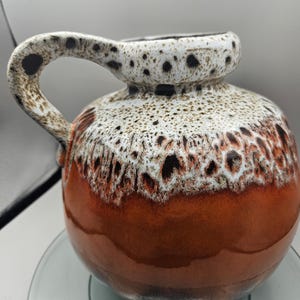 以下が含まれることがあります： 茶色、白、黒の斑点模様の釉薬が施されたセラミックの花瓶。花瓶は丸いボディとシングルハンドルです。