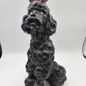 May include: Black poodle figurine with a red bow, sitting with its head tilted up. The figurine is made of a textured material that resembles fur.