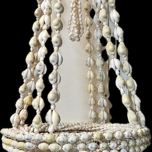 Könnte beinhalten: Ein handgefertigter Kronleuchter aus verschiedenen Muscheln, der eine natürliche, cremefarbene Farbpalette aufweist. Das Design weist mehrere Stränge aus Muscheln auf, die einen Kaskadeneffekt erzeugen. Der Kronleuchter ist an einem kreisförmigen Rahmen aufgehängt.