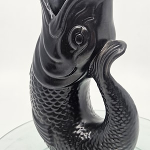 May include: A black ceramic vase in the shape of a fish. The vase has a wide opening and a curved tail. The fish's scales are textured, and the vase sits on a small, round base. The vase is a decorative item.