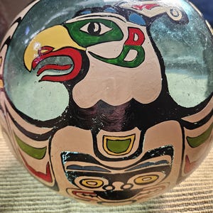 May include: A round, glass bauble with a painted design of a Native American eagle in green, red, yellow and black.