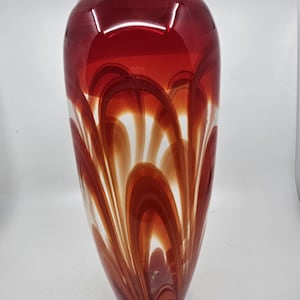 May include: A tall, decorative glass vase with a rounded top and a flared base. The vase features a vibrant color scheme, with a deep red top that transitions into a pattern of red, orange, and white swirls. The vase is approximately 12 inches tall.