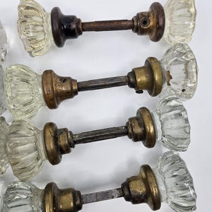 Peut inclure: Ensemble de boutons de porte anciens en verre avec des garnitures en laiton et marron foncé. Les boutons en verre transparent présentent un motif côtelé. La quincaillerie comprend des tiges filetées et des colliers en laiton. Les boutons sont disposés sur fond blanc.