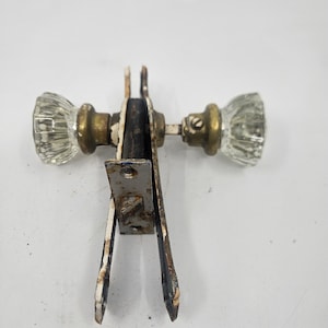 Peut inclure: Un ensemble de quincaillerie de porte vintage comprenant deux boutons en verre transparent à facettes fixés sur des tiges en métal doré. Le mécanisme central est un loquet rectangulaire en métal avec une finition blanche écaillée et usée.